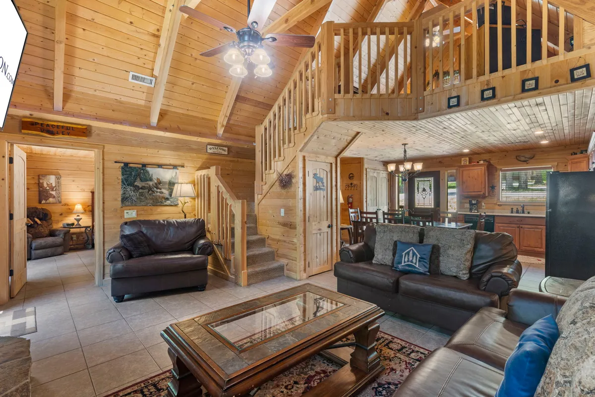 This view encompasses both a cozy seating area and a dining table adjacent to a prominent stone fireplace, set beneath rustic wooden beams. The arrangement highlights how the kitchen, dining, and living areas are interconnected, all within a warmly lit, inviting ambiance.