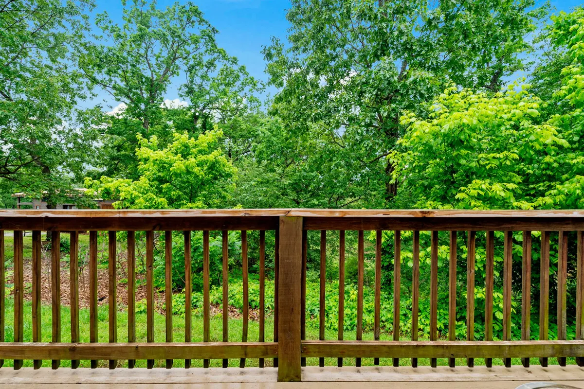 A tranquil view from a wooden deck, surrounded by lush green trees and an open sky. The wooden railing emphasizes the cabin’s rustic charm, and the setting is perfect for relaxing with a loved one while taking in the peaceful natural surroundings.