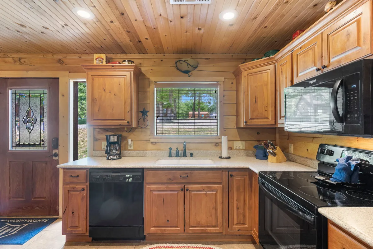A bright, fully equipped kitchen is showcased here, featuring wooden cabinets, modern black appliances, a window view, and well-organized countertops adorned with decorative touches. The scene conveys the space’s readiness for cooking and entertaining, while still feeling homey and inviting.