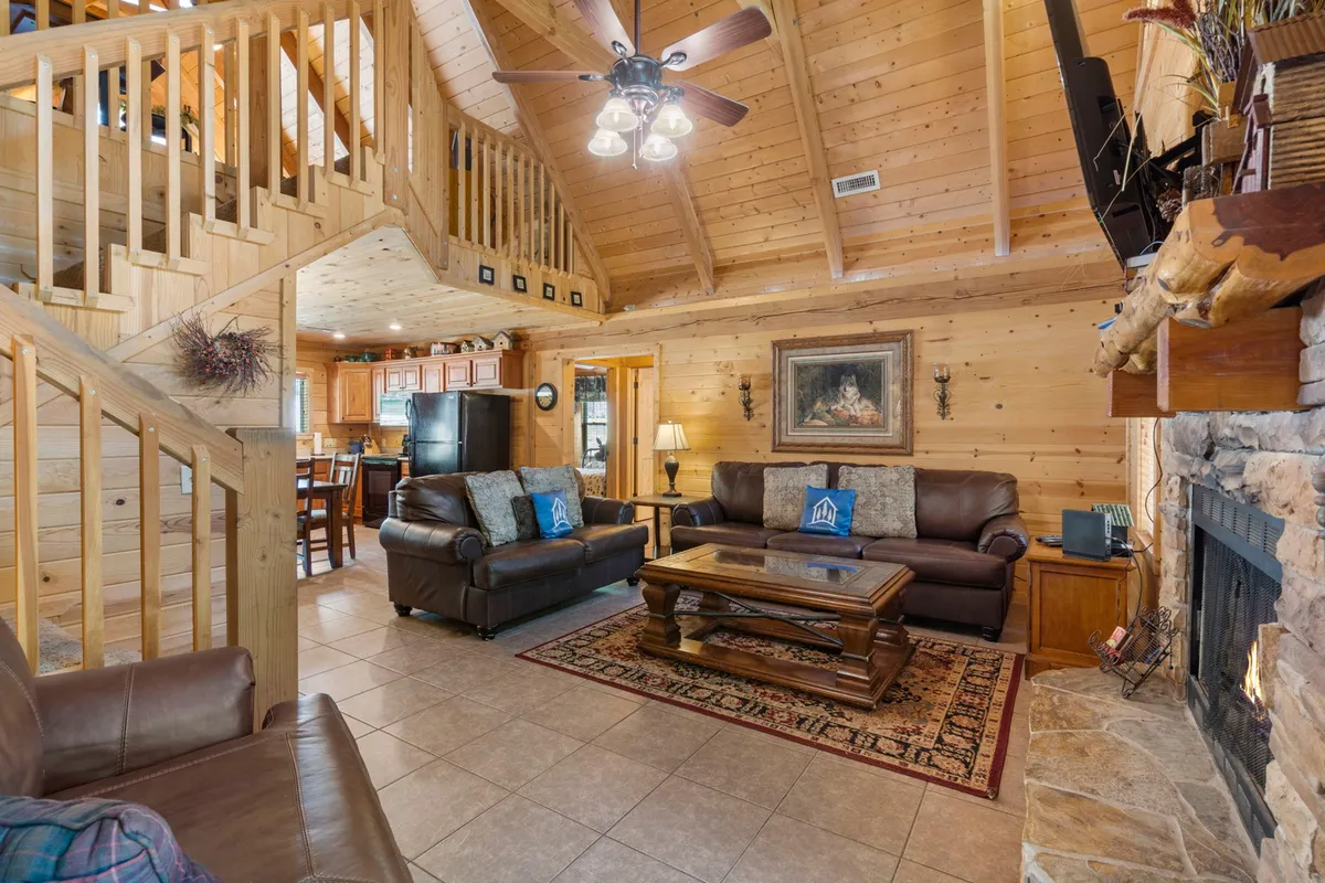 This image captures a spacious great room with large wooden beams, a striking stone fireplace, and comfortable seating arranged for conversation or relaxation. The adjacent dining space is visible, showing how seamlessly the living and dining areas flow together. The mood exudes warmth, making it ideal for gatherings or cozy evenings by the fire.