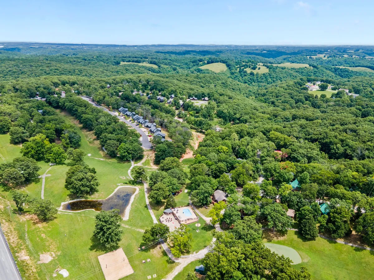An expansive aerial view captures a cluster of rustic cabins nestled among rolling green hills, with a sparkling swimming pool and pond in the foreground. Recreational areas and winding trails are visible, inviting guests to hike, swim, and explore the outdoors with family.