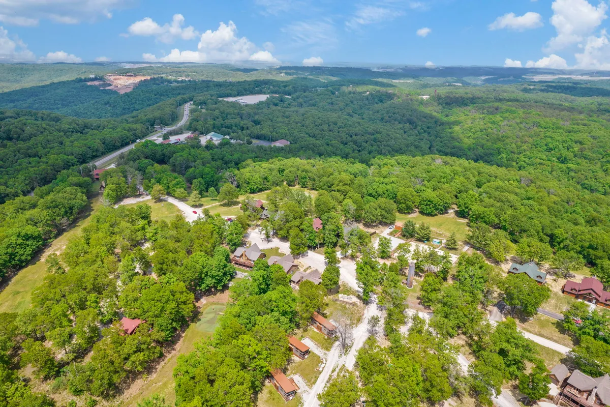 A wide view of multiple log cabins nestled in lush greenery, with winding paths connecting the properties. Distant hills and a tranquil atmosphere invite exploration and adventure with pets.