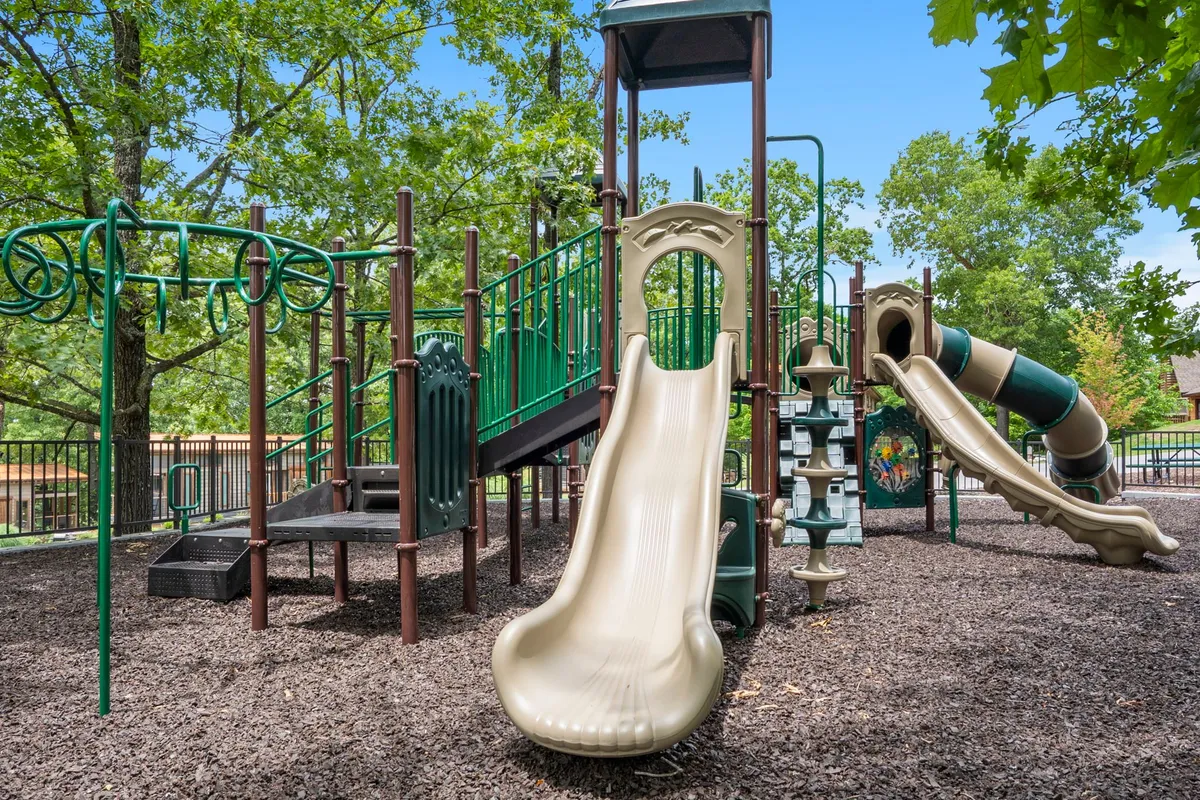 A colorful playground with slides and climbing structures sits among green grass and trees, as children run and play while parents supervise in the background. The joyful mood showcases opportunities for outdoor family bonding and playdates.