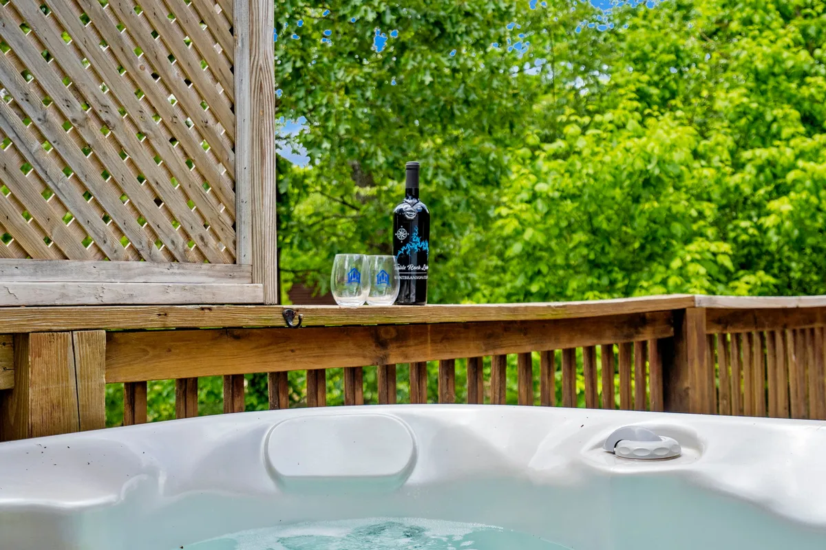 A steaming hot tub set on a private deck, with a wine bottle and glasses close by, looking out over natural surroundings. The environment is tranquil, suggesting relaxation after a day of outdoor activities.