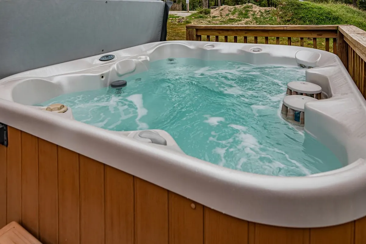 An inviting outdoor hot tub sits on a private deck surrounded by natural landscape, offering views into the trees and a tranquil atmosphere for relaxation. The deck is spacious, providing room for socializing and enjoying the outdoors, whether by day or under the stars.