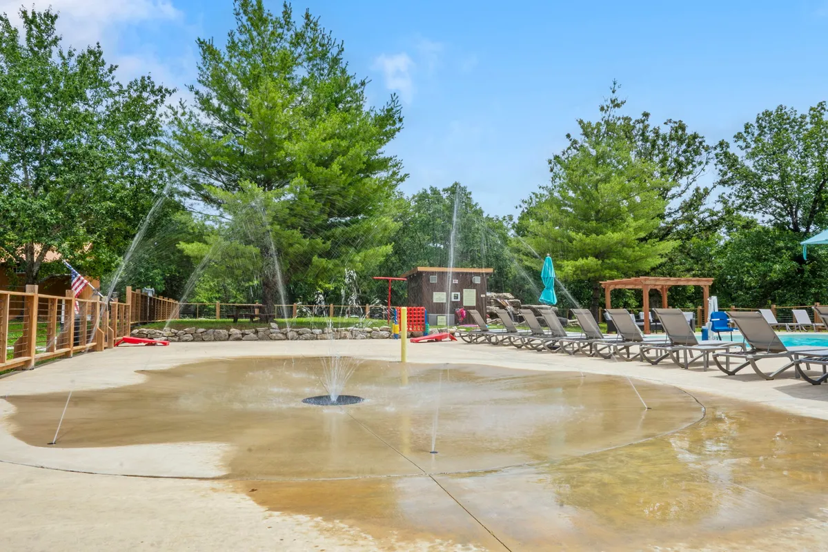 A lively splash pad and pool area surrounded by lounge chairs, with children playing and families gathering under the warm sun. Water fountains and a playful atmosphere dominate the scene, evoking laughter and summer excitement.