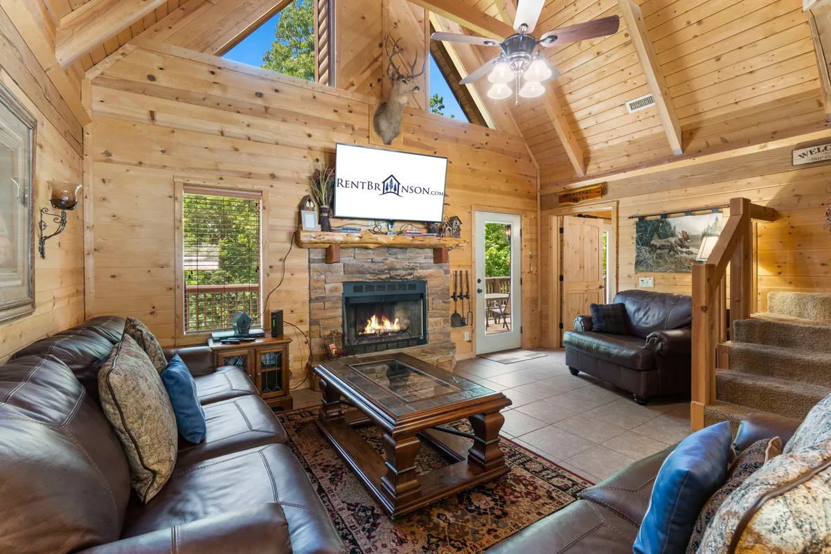 The hero image showcases a spacious, light-filled living room with classic log cabin architecture, including exposed wooden beams and a cozy stone fireplace as the centerpiece. A plush sofa invites relaxation, while a large TV and soft rug suggest a perfect spot for movie nights or gathering with family and friends. The warm wood tones and comfortable furnishings create an inviting, homey atmosphere ideal for unwinding after a day of adventure.