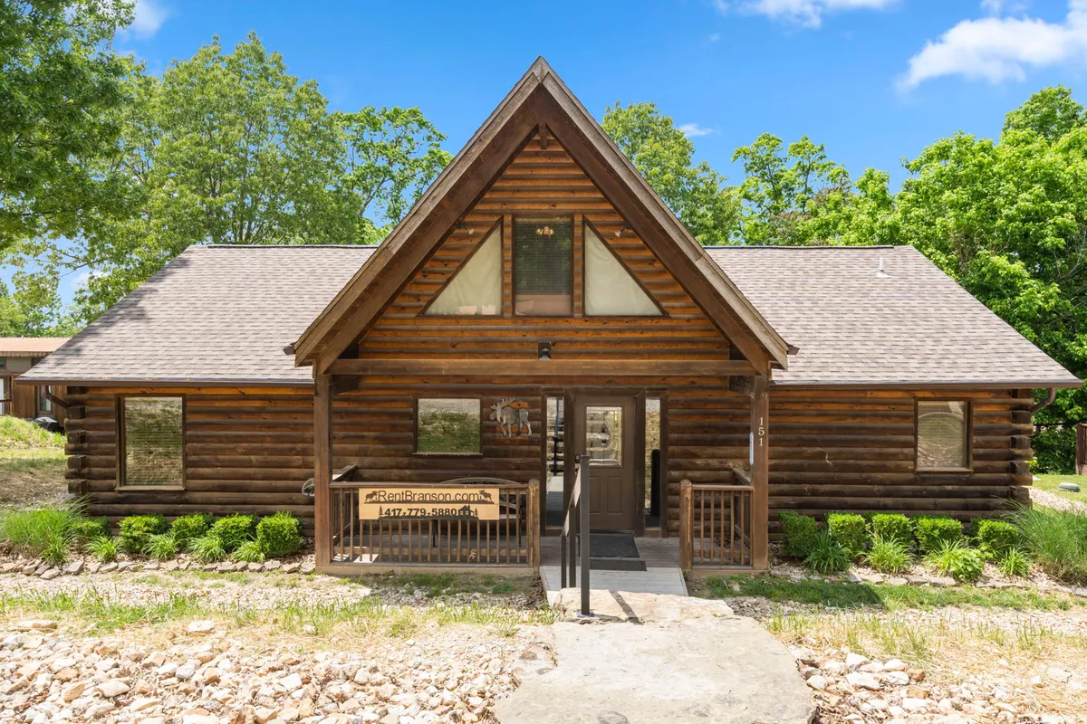 A full view of the log cabin’s exterior, featuring prominent large front windows, wooden steps, and a shaded deck area. Surrounded by greenery, the space is ideal for shared moments outdoors under the stars.