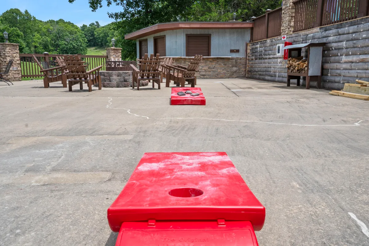 A cozy outdoor fire pit area features red cornhole boards and wooden chairs, inviting guests to gather for games and fireside chats as the sun sets. The landscape beyond hints at wide open spaces and the potential for creating lasting memories around the fire.