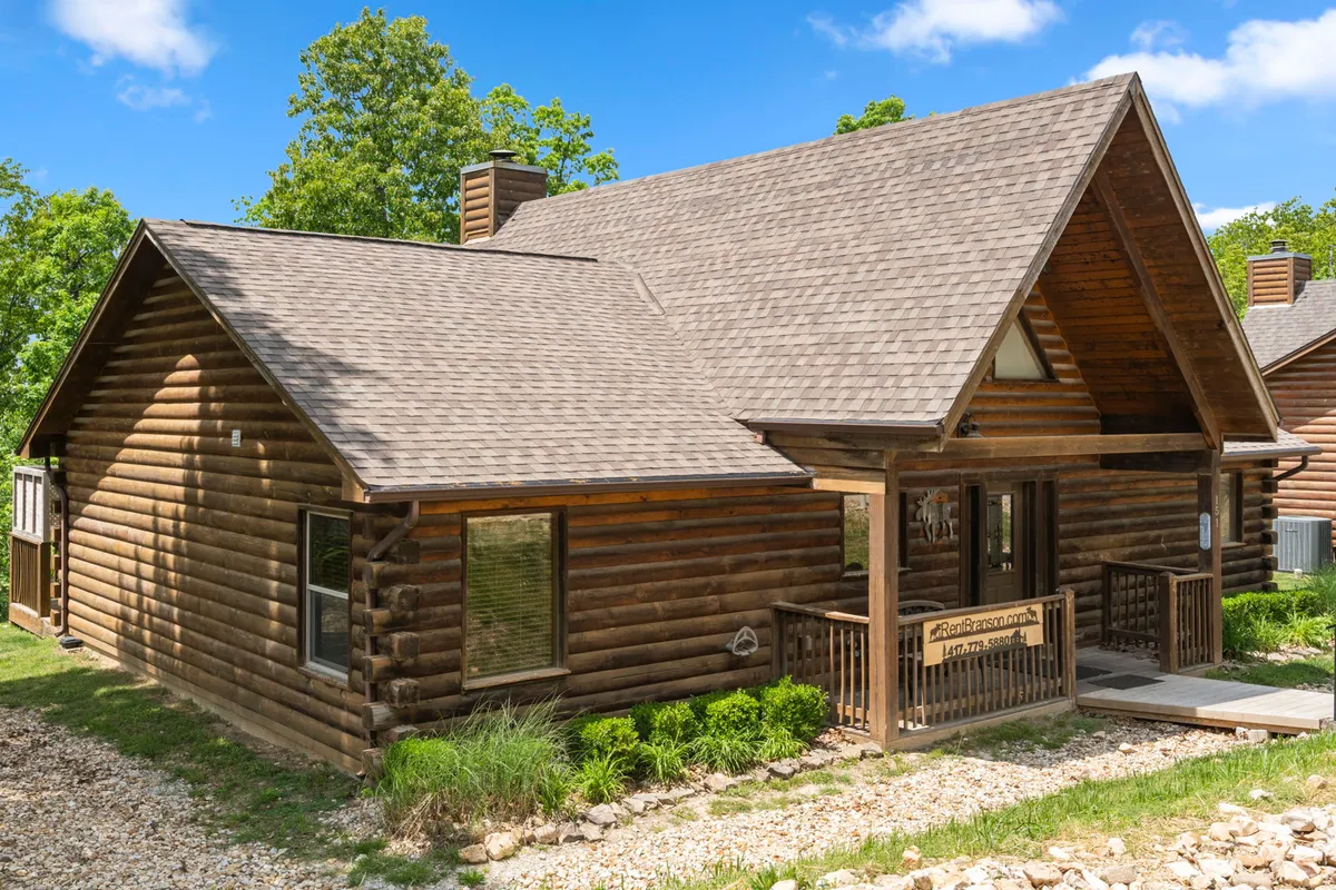 A charming log cabin with a welcoming front porch set amid mature trees and a landscaped area. The porch features seating that invites guests to relax and enjoy the outdoors, while paths and greenery hint at nearby walking routes.