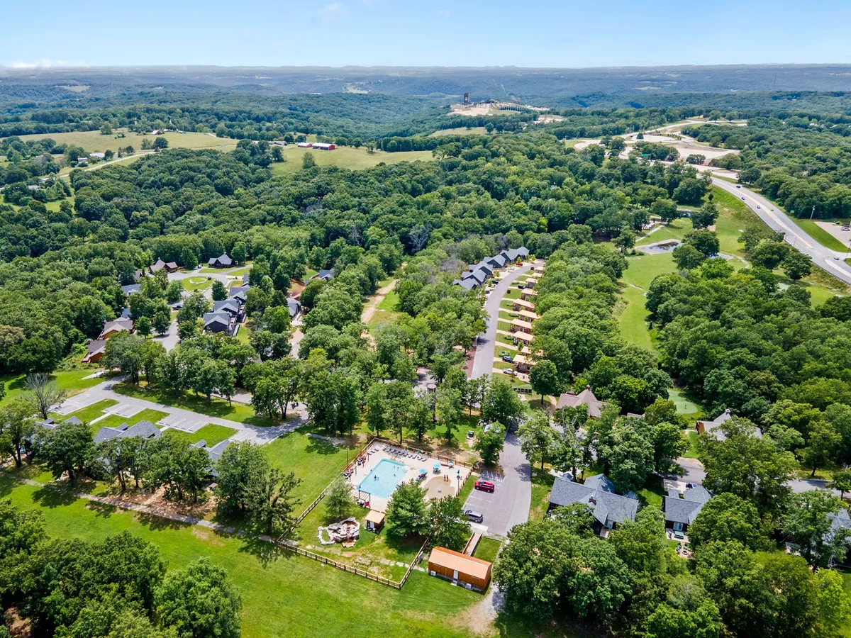 A vibrant community scene featuring a swimming pool surrounded by lounge chairs, with a cluster of inviting cabins set amidst a lush, expansive green landscape. The setting suggests opportunities for swimming, picnicking, hiking, and gathering, with pathways connecting different areas of the resort.