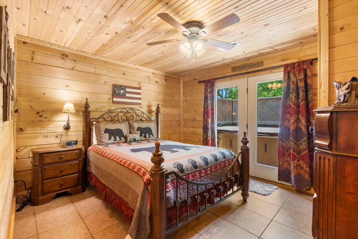 A cozy bedroom featuring a bed with bear-themed bedding, warm wooden walls and ceiling, draped curtains, and a lamp on the bedside table. Natural light streams into the room, creating a restful and inviting atmosphere ideal for relaxation.