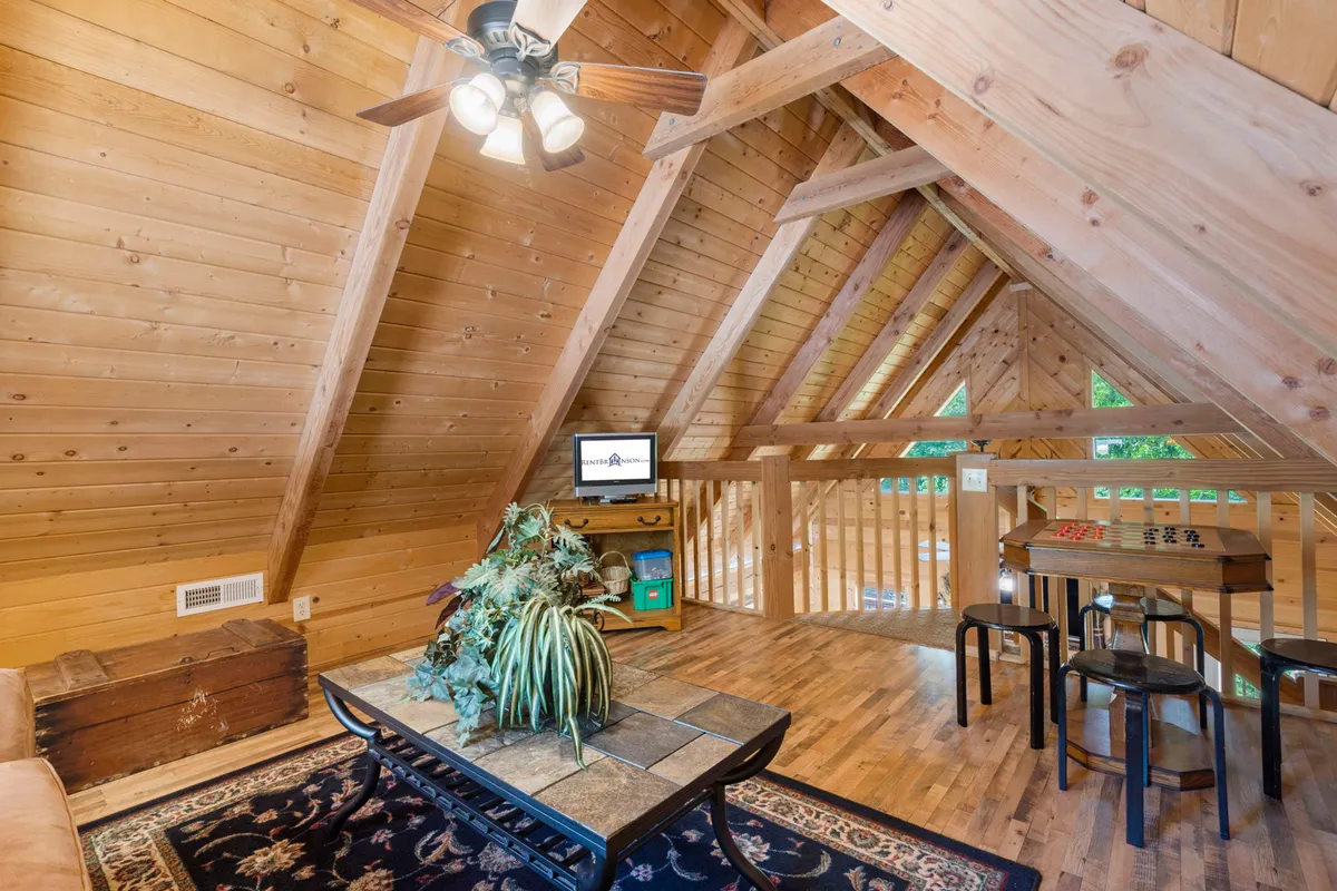 A cozy loft space featuring wooden beams, a stone-topped coffee table set for games, a TV, and a multi-purpose game table. The area feels warm and inviting, ideal for game nights, movie marathons, and friendly gatherings away from the main living area.